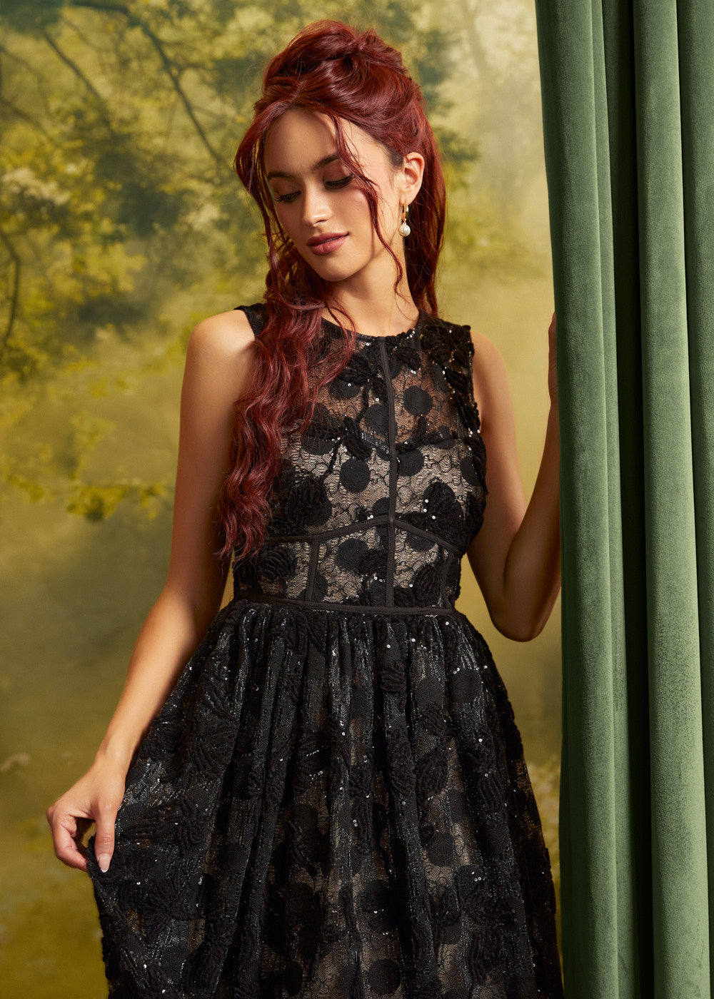 A woman with long red hair wearing a sleeveless black lace formal dress with a sheer bodice and flared skirt, posed beside a green curtain against a soft green backdrop.