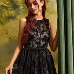 A woman with long red hair wearing a sleeveless black lace formal dress with a sheer bodice and flared skirt, posed beside a green curtain against a soft green backdrop.