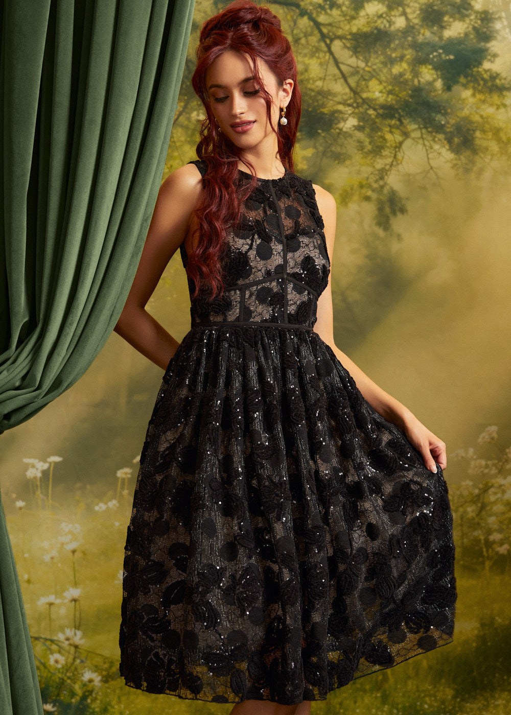 A woman wearing a sleeveless black lace dress with a fitted waist and full skirt, posed in front of a soft-focus green and yellow outdoor backdrop with a curtain on the left.
