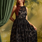A woman wearing a sleeveless black lace dress with a fitted waist and full skirt, posed in front of a soft-focus green and yellow outdoor backdrop with a curtain on the left.