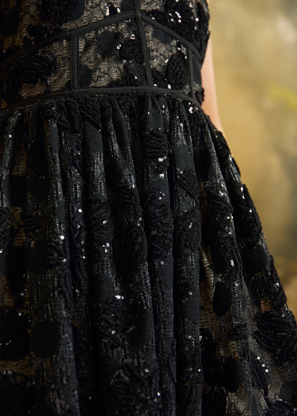 Close-up of a black embellished lace dress with sequins and textured floral embroidery, showing the bodice and gathered skirt against a softly blurred warm-toned background.