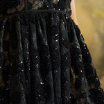 Close-up of a black embellished lace dress with sequins and textured floral embroidery, showing the bodice and gathered skirt against a softly blurred warm-toned background.