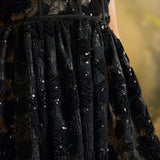 Close-up of a black embellished lace dress with sequins and textured floral embroidery, showing the bodice and gathered skirt against a softly blurred warm-toned background.