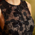 Close-up of a cocktail dress's round neckline and sheer black floral lace bodice with dimensional embroidery, shimmering sequins, and decorative seam detailing.