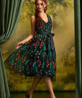 A woman models a sleeveless V-neck midi dress with a dark green and black floral print, a black waist tie, and black pointed-toe heels, posed in front of a scenic forest backdrop with green curtains.