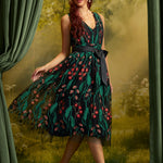 A woman models a sleeveless V-neck midi dress with a dark green and black floral print, a black waist tie, and black pointed-toe heels, posed in front of a scenic forest backdrop with green curtains.