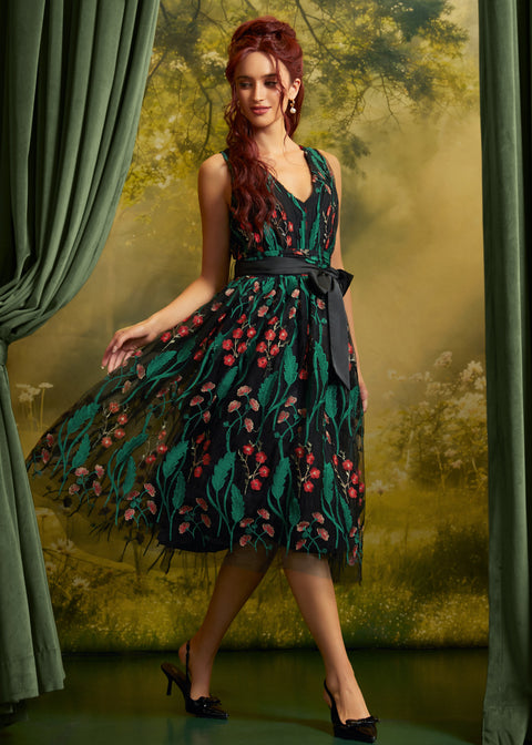 A woman models a sleeveless V-neck midi dress with a dark green and black floral print, a black waist tie, and black pointed-toe heels, posed in front of a scenic forest backdrop with green curtains.