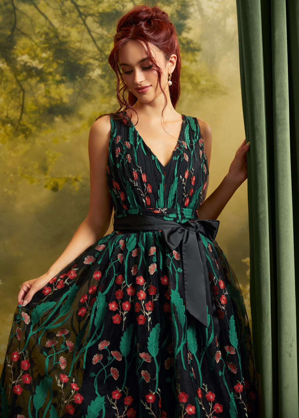 A woman wearing a sleeveless V-neck ball gown with a green and black floral vine pattern and a matching black waist sash, posed next to a green curtain.