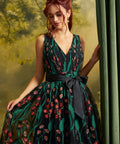 A woman wearing a sleeveless V-neck ball gown with a green and black floral vine pattern and a matching black waist sash, posed next to a green curtain.