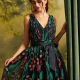 A woman wearing a sleeveless V-neck ball gown with a green and black floral vine pattern and a matching black waist sash, posed next to a green curtain.