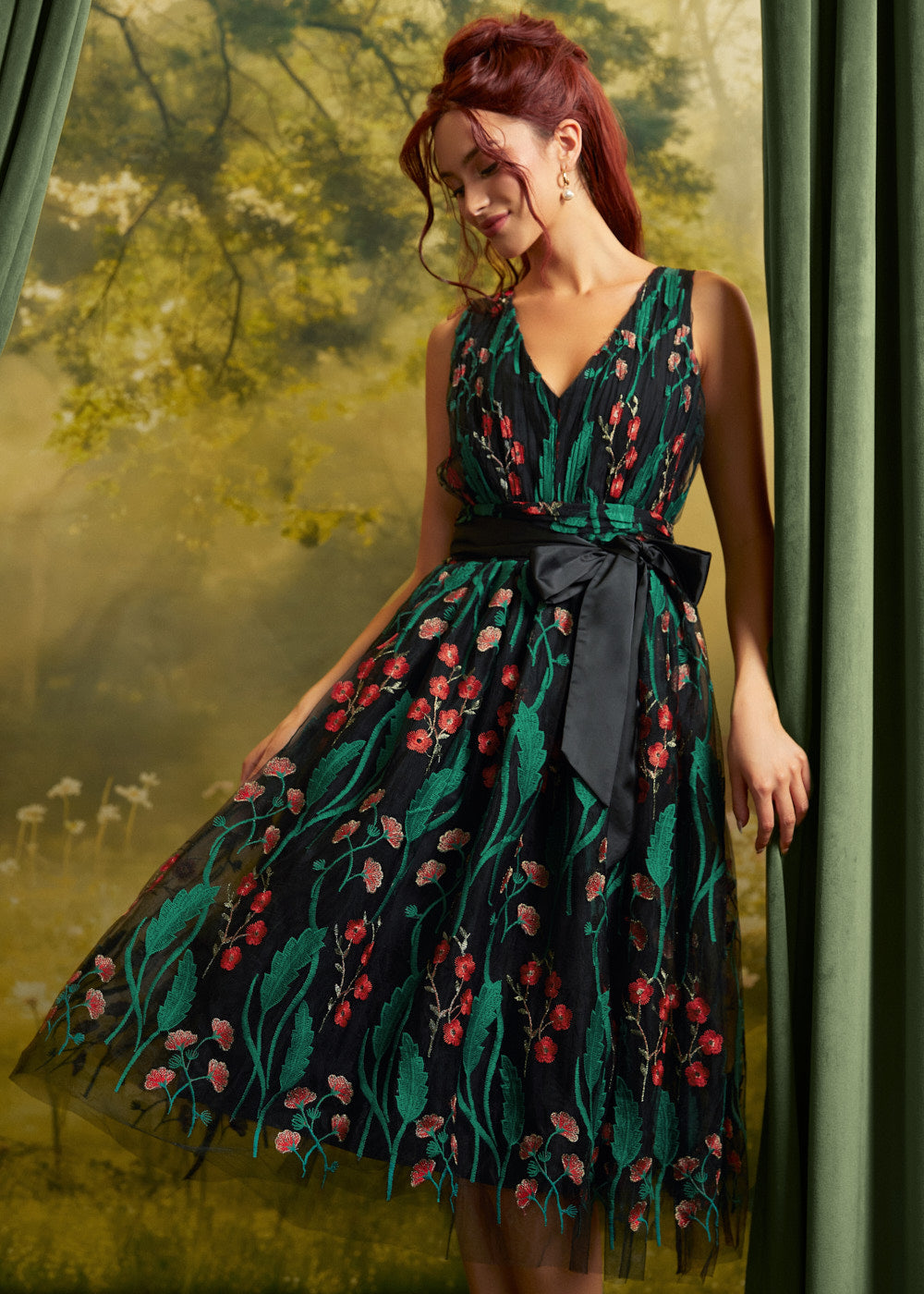 A woman models a sleeveless V-neck midi dress with a dark green and black floral print, featuring a black waist tie belt. She is posed in front of a soft-focus green forest backdrop with draped curtains on the sides.