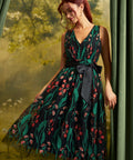 A woman models a sleeveless V-neck midi dress with a dark green and black floral print, featuring a black waist tie belt. She is posed in front of a soft-focus green forest backdrop with draped curtains on the sides.
