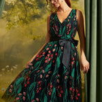 A woman models a sleeveless V-neck midi dress with a dark green and black floral print, featuring a black waist tie belt. She is posed in front of a soft-focus green forest backdrop with draped curtains on the sides.