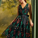A woman models a sleeveless V-neck midi dress with a dark green and black floral print, featuring a black waist tie belt. She is posed in front of a soft-focus green forest backdrop with draped curtains on the sides.