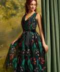 A woman models a sleeveless V-neck tulle dress with a black waist sash, featuring a dark green and black floral print with red and pink blossoms, posed against a green curtain and a soft-focus forest backdrop.