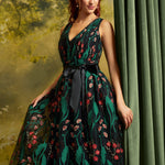 A woman models a sleeveless V-neck tulle dress with a black waist sash, featuring a dark green and black floral print with red and pink blossoms, posed against a green curtain and a soft-focus forest backdrop.