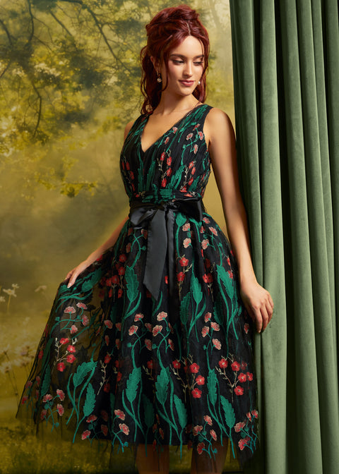 A woman models a sleeveless V-neck tulle dress with a black waist sash, featuring a dark green and black floral print with red and pink blossoms, posed against a green curtain and a soft-focus forest backdrop.