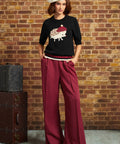 A woman stands against a brick wall wearing a black knit top with a hedgehog graphic, deep burgundy wide-leg trousers, and a cream beret. A vintage dark brown trunk sits to the left.