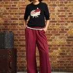A woman stands against a brick wall wearing a black knit top with a hedgehog graphic, deep burgundy wide-leg trousers, and a cream beret. A vintage dark brown trunk sits to the left.