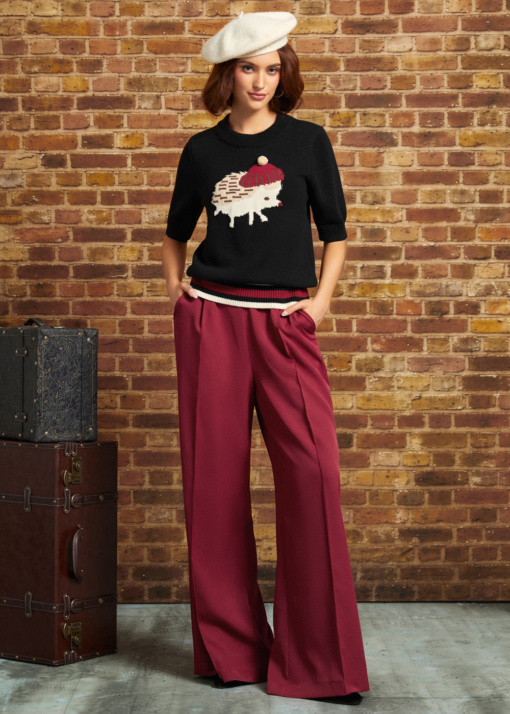 A woman stands against a brick wall wearing a black knit top with a hedgehog graphic, deep burgundy wide-leg trousers, and a cream beret. A vintage dark brown trunk sits to the left.