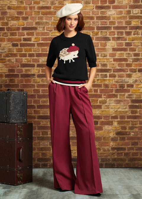 A woman stands against a brick wall wearing a black knit top with a hedgehog graphic, deep burgundy wide-leg trousers, and a cream beret. A vintage dark brown trunk sits to the left.