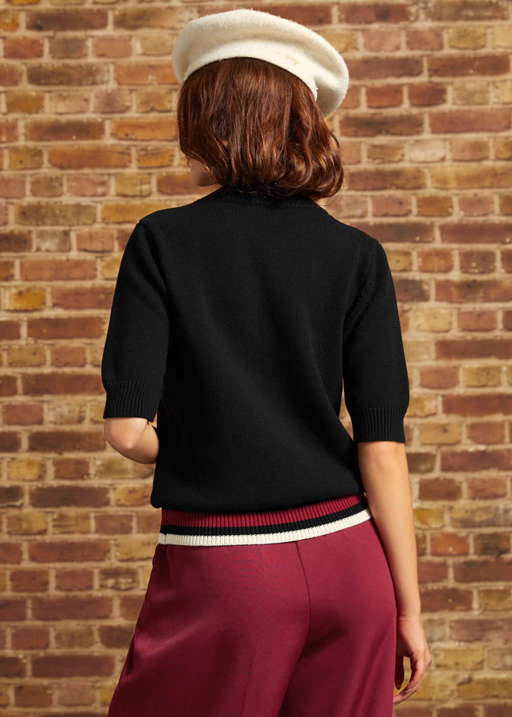 Back view of the black cotton Hattie Half-Sleeve Sweater showing the plain knit back, ribbed short sleeve cuffs, and contrasting burgundy, black, and cream striped hem.