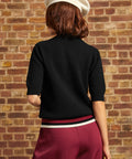 Back view of the black cotton Hattie Half-Sleeve Sweater showing the plain knit back, ribbed short sleeve cuffs, and contrasting burgundy, black, and cream striped hem.