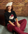 A woman sits on a concrete floor against a brick wall, wearing a cream beret, a black knit top with a hedgehog graphic, and burgundy wide-leg trousers.