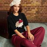 A woman sits on a concrete floor against a brick wall, wearing a cream beret, a black knit top with a hedgehog graphic, and burgundy wide-leg trousers.