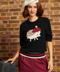 A woman models a black knit sweater featuring a knitted hedgehog design with a red beret and cream color blocking, styled with burgundy trousers and a cream beret against a brick wall background.