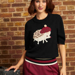 A woman models a black knit sweater featuring a knitted hedgehog design with a red beret and cream color blocking, styled with burgundy trousers and a cream beret against a brick wall background.