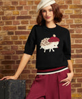 A woman models a black knit sweater featuring a white hedgehog wearing a red beret, paired with burgundy wide-leg trousers and a cream beret, posed against a rustic brick wall.