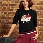 A woman models a black knit sweater featuring a white hedgehog wearing a red beret, paired with burgundy wide-leg trousers and a cream beret, posed against a rustic brick wall.
