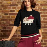 A woman models a black knit sweater featuring a white hedgehog wearing a red beret, paired with burgundy wide-leg trousers and a cream beret, posed against a rustic brick wall.