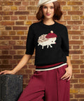 A woman wearing a black knit sweater with a hedgehog graphic, burgundy wide-leg pants, and a cream beret, posing against a brick wall next to a vintage suitcase.
