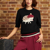 A woman wearing a black knit sweater with a hedgehog graphic, burgundy wide-leg pants, and a cream beret, posing against a brick wall next to a vintage suitcase.