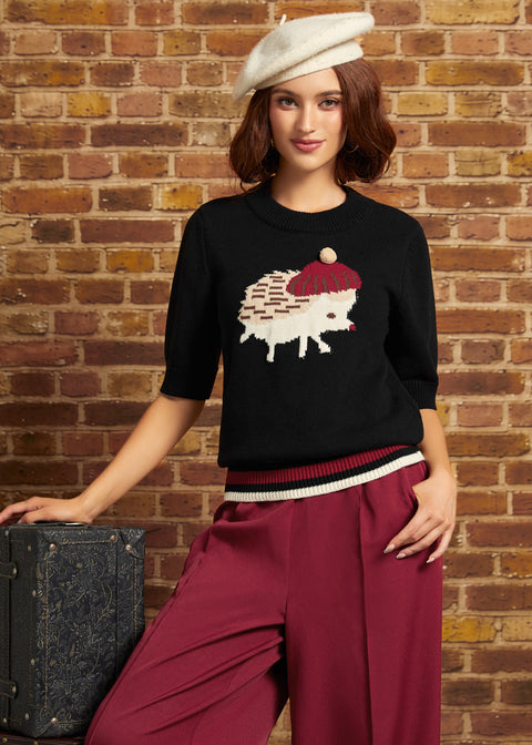 A woman wearing a black knit sweater with a hedgehog graphic, burgundy wide-leg pants, and a cream beret, posing against a brick wall next to a vintage suitcase.