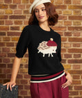 Model wearing the black hedgehog novelty sweater with a ribbed crew neckline, elbow-length sleeves, burgundy and cream striped hem, and hedgehog motif on the chest, paired with burgundy trousers.