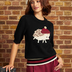 Model wearing the black hedgehog novelty sweater with a ribbed crew neckline, elbow-length sleeves, burgundy and cream striped hem, and hedgehog motif on the chest, paired with burgundy trousers.