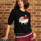 Model wearing the black hedgehog novelty sweater with a ribbed crew neckline, elbow-length sleeves, burgundy and cream striped hem, and hedgehog motif on the chest, paired with burgundy trousers.