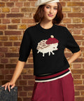 A woman wearing a black knit sweater with a knitted hedgehog graphic, a white beret, and burgundy pants, posing against a brick wall.