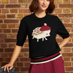 A woman wearing a black knit sweater with a knitted hedgehog graphic, a white beret, and burgundy pants, posing against a brick wall.