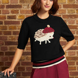 A woman wearing a black knit sweater with a knitted hedgehog graphic, a white beret, and burgundy pants, posing against a brick wall.