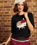 Model wearing the black ModCloth Hattie Half-Sleeve Sweater featuring a cream hedgehog in a burgundy beret with a pom-pom, striped ribbed hem, and elbow-length sleeves, styled with burgundy trousers and a cream beret.