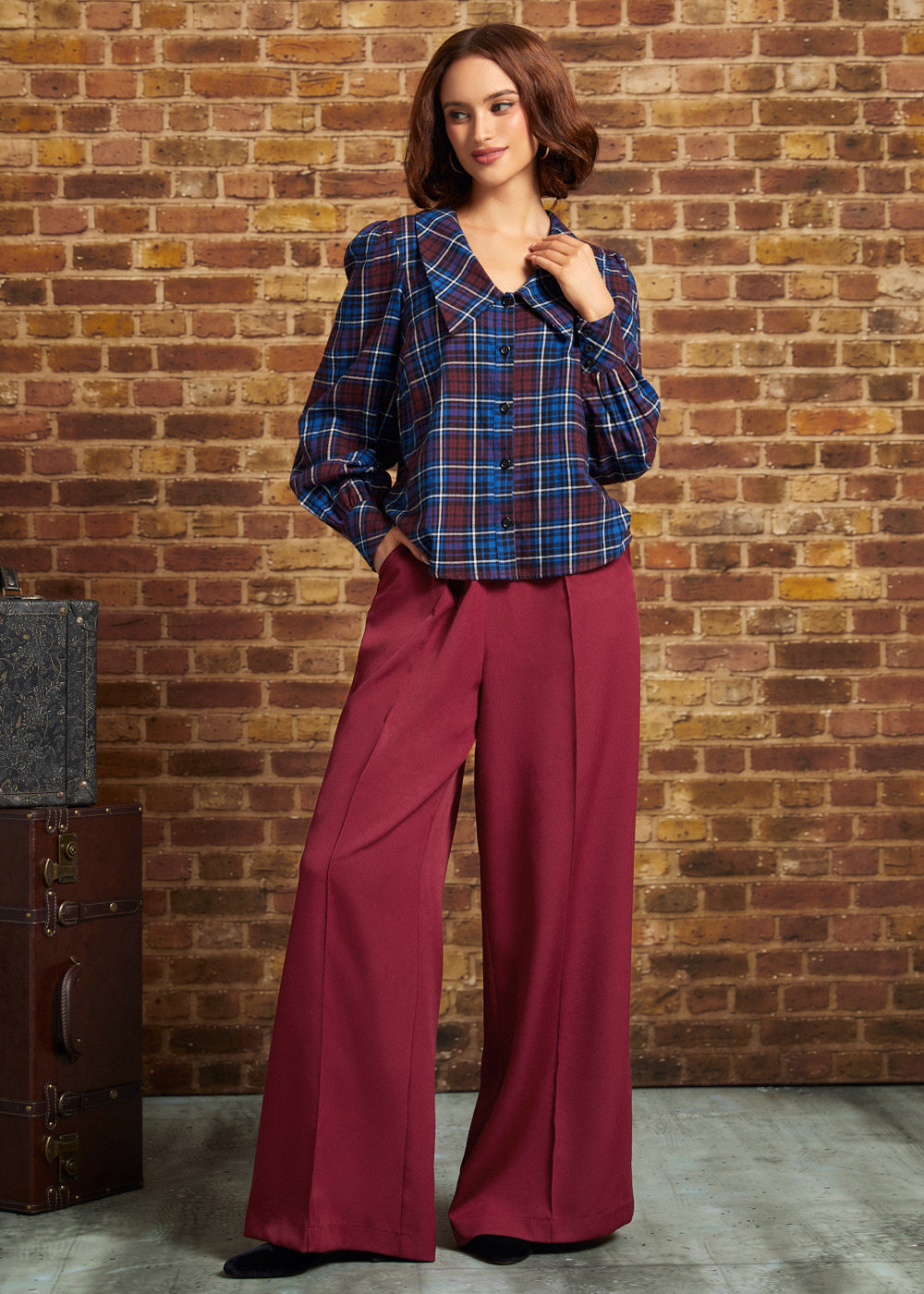 A woman stands in front of a brick wall wearing a dark blue and red plaid button-up shirt with voluminous sleeves and deep burgundy wide-leg trousers.