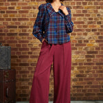 A woman stands in front of a brick wall wearing a dark blue and red plaid button-up shirt with voluminous sleeves and deep burgundy wide-leg trousers.