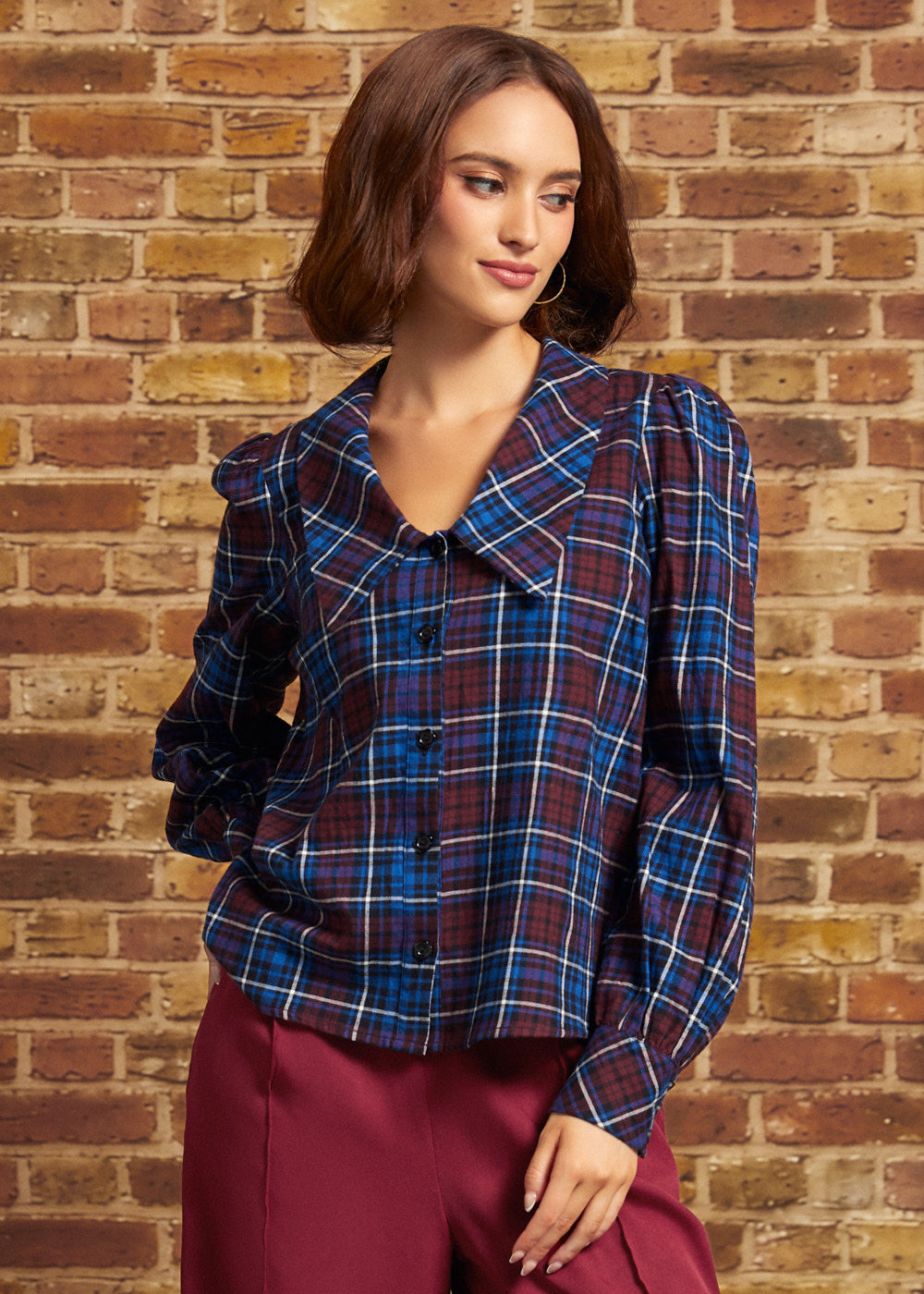 Model wearing a burgundy and blue plaid cotton blouse with oversized pointed collar, long puff sleeves, and black button-front closure, paired with burgundy trousers against a brick wall.