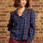 Model wearing a burgundy and blue plaid cotton blouse with oversized pointed collar, long puff sleeves, and black button-front closure, paired with burgundy trousers against a brick wall.