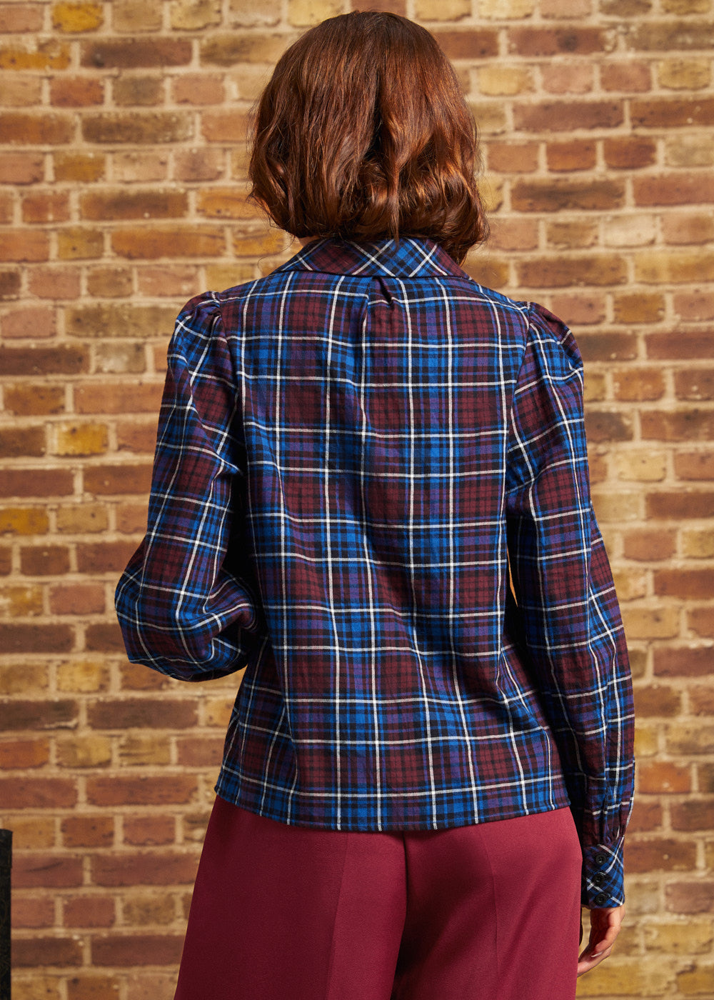 Rear view of a burgundy and blue plaid long sleeve blouse showing gathered shoulder seams, relaxed fit, and buttoned cuffs against a brick wall.