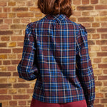 Rear view of a burgundy and blue plaid long sleeve blouse showing gathered shoulder seams, relaxed fit, and buttoned cuffs against a brick wall.